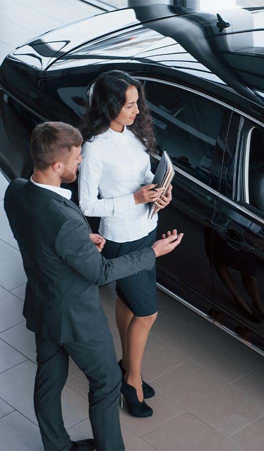Leasing versus Buying a Chevrolet at Delaney Chevrolet of Greensburg in Greensburg, PA | Sales woman holding documents while showing customer in suit some car options on the sales floor