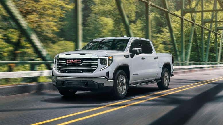 A white 2026 GMC Sierra 1500 driving down the road over a bridge by a forest.