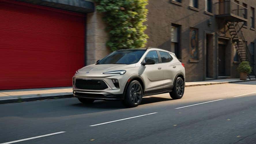 A beige Buick Encore GX driving down the road through a city.