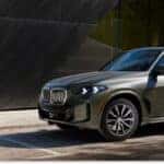 Front three-quarter view of a dark green 2026 BMW X5 parked outside a modern building.