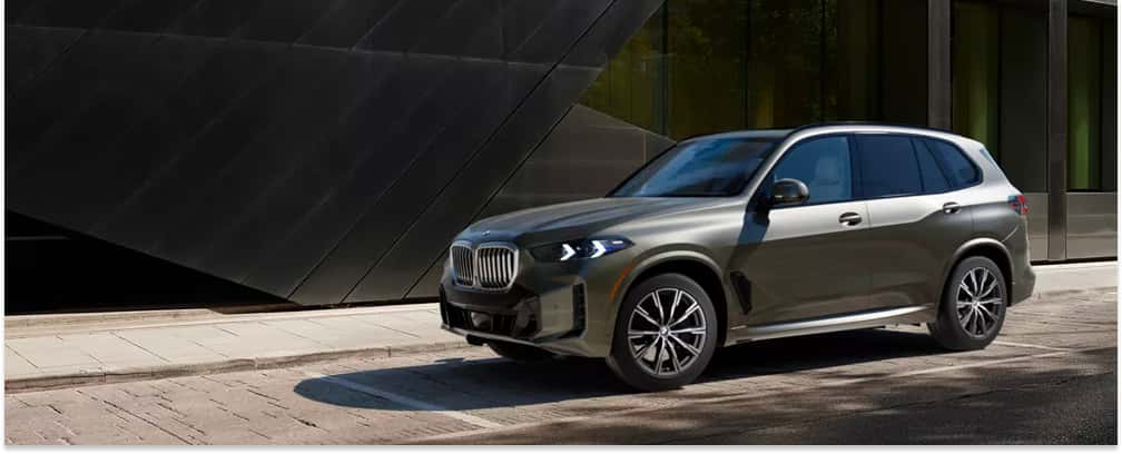 Front three-quarter view of a dark green 2026 BMW X5 parked outside a modern building.
