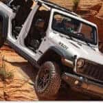 > Front three-quarter view of a white 2026 Jeep Wrangler with its doors and top removed, navigating a steep, rugged incline on red sandstone rock formations.