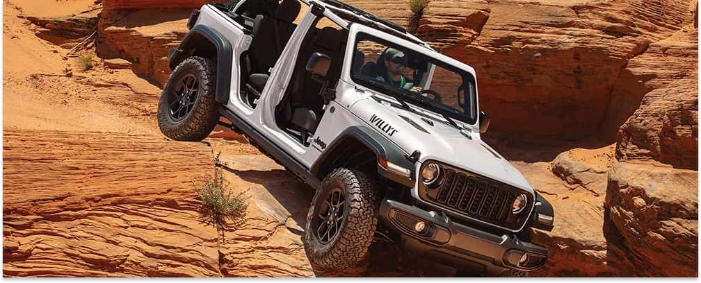 > Front three-quarter view of a white 2026 Jeep Wrangler with its doors and top removed, navigating a steep, rugged incline on red sandstone rock formations.