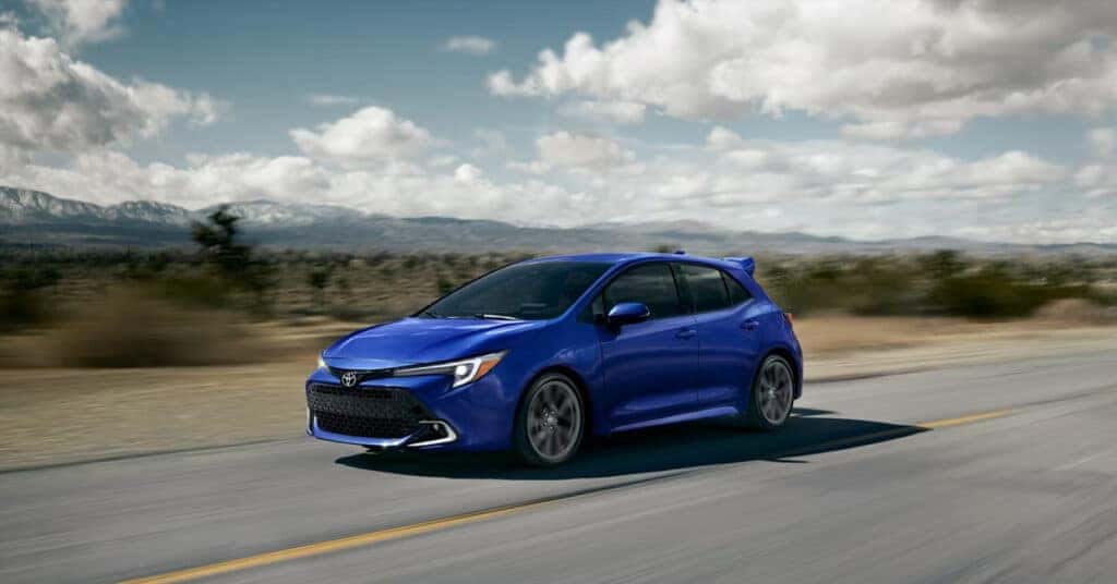2026 Toyota Corolla Hatchback driving on a scenic road with mountains in the background.